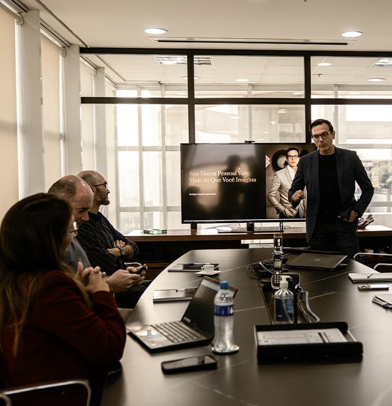 Rodrigo Baraldi, de pé, conduz um encontro com advogados reunidos em volta de uma mesa, diante de uma tela de apresentação